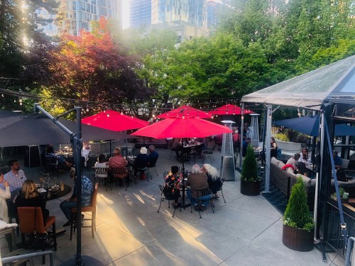 Outdoor dining area with red umbrellas, surrounded by greenery and people enjoying their meals in a lively atmosphere.