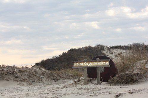 Sign for False Cape Landing on a sandy beach with dunes and cloudy sky in the background.