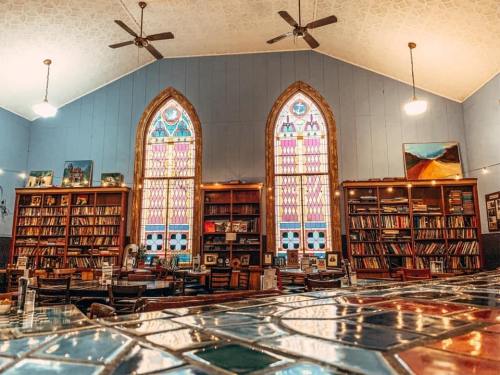 Cozy library with tall stained glass windows, wooden bookshelves filled with books, and a reflective table surface.