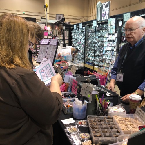 A vendor assists a customer at a craft fair booth filled with colorful supplies and products.