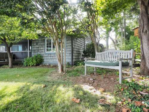 A wooden bench sits in a grassy area surrounded by trees, with a house visible in the background.