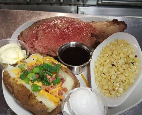 A plate featuring a juicy prime rib, baked potato with toppings, corn, and sides of butter and sauce.