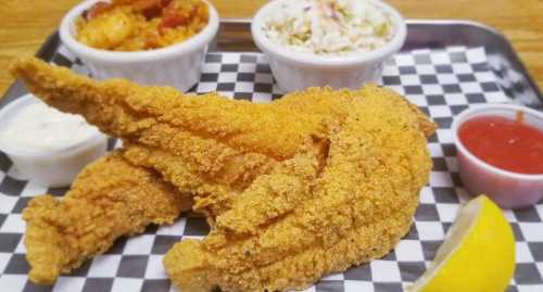 A plate of golden fried fish fillets with coleslaw, shrimp, and dipping sauces on a checkered tray.