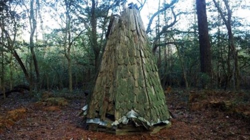 A small, weathered wooden structure resembling a teepee, surrounded by trees in a forested area.