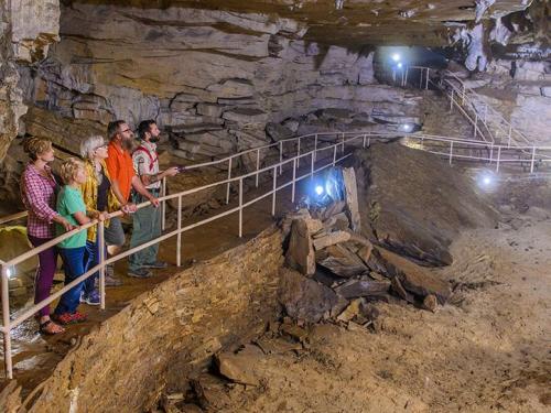 A group of people exploring a cave, standing on a path with rock formations and lights illuminating the surroundings.