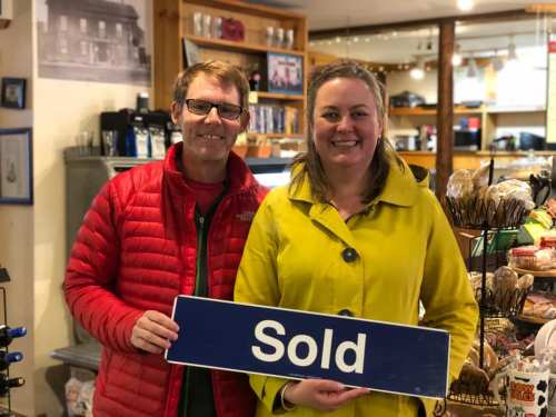 Two smiling individuals hold a "Sold" sign in a cozy shop setting.