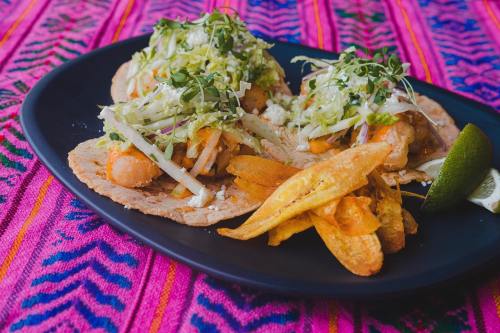 Three tacos topped with lettuce and herbs, served with plantain chips and a lime wedge on a colorful textile background.