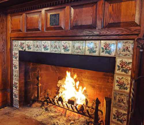 A cozy fireplace with a roaring fire, surrounded by decorative tiles and wooden paneling.