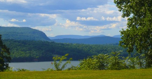 Scenic view of a lake surrounded by lush green hills and mountains under a partly cloudy sky.