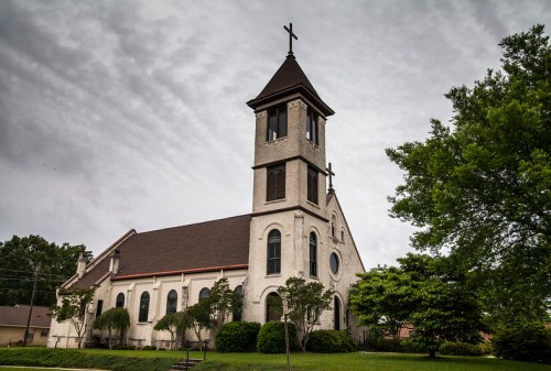 A tall church with a steeple and cross, surrounded by trees under a cloudy sky.