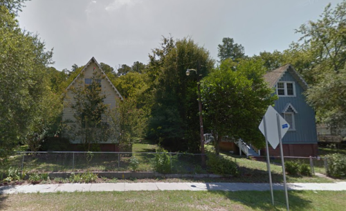 Two houses with steep roofs surrounded by trees, set behind a fence, in a green, wooded area.