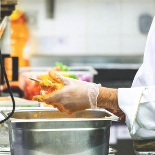 A person wearing gloves prepares food in a kitchen, with pots and ingredients visible in the background.
