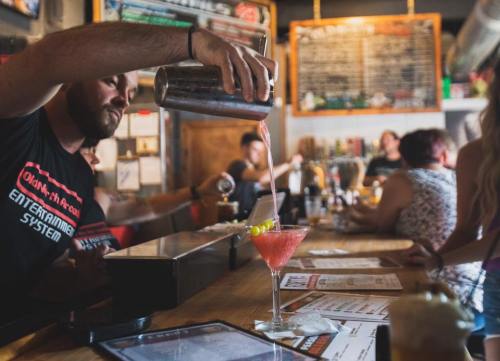 A bartender pours a cocktail into a glass at a lively bar with patrons in the background.