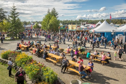 A bustling outdoor festival scene with crowds, food stalls, and picnic tables surrounded by flowers and trees.