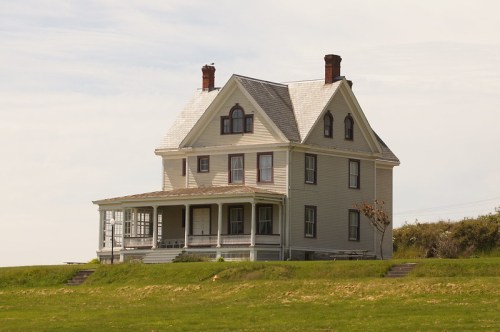 A large, two-story Victorian-style house with a porch, set on a grassy landscape under a clear sky.