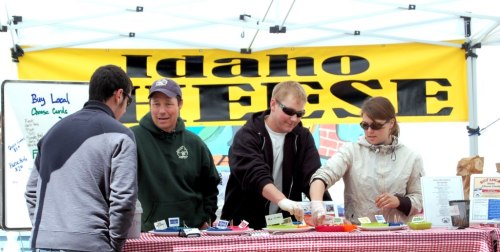 A group of people at a market booth labeled "Idaho Cheese," showcasing various cheese products and samples.