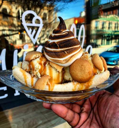 A hand holds a glass bowl filled with a banana split topped with toasted meringue, cookies, and caramel drizzle.