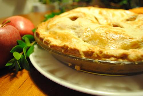 A golden-brown apple pie in a glass dish, with fresh apples and greenery nearby on a wooden table.