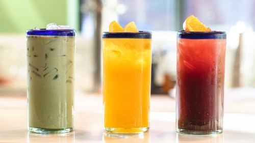 Three colorful drinks in tall glasses: green iced drink, orange juice, and a red beverage, each garnished with orange slices.