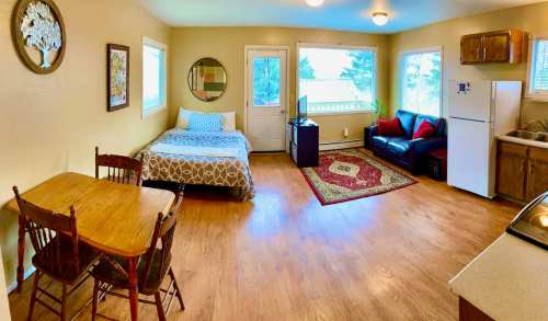 Cozy studio apartment with a bed, dining table, couch, TV, and kitchen area, featuring large windows and warm decor.