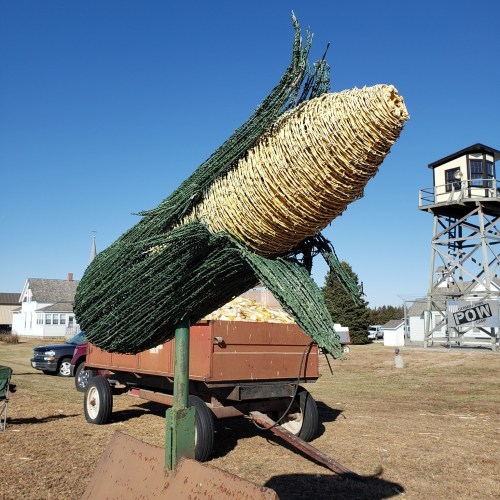 A large corn sculpture stands on a post, with a trailer in the background and a watchtower in the distance.