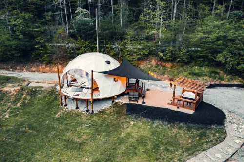Aerial view of a unique dome-shaped cabin surrounded by greenery, featuring a patio and shaded seating area.