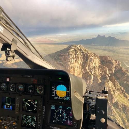 Cockpit view of a helicopter flying over rugged mountains and valleys under a cloudy sky.