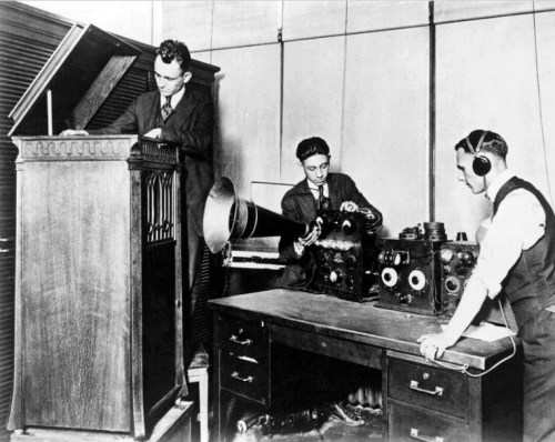 Three men in early 20th-century attire work with vintage audio equipment in a studio setting.