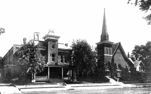 Historic building with a tall spire, surrounded by trees, featuring a porch and a church structure beside it.