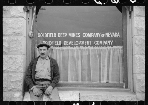 A man sits outside a window displaying "Goldfield Deep Mines Company of Nevada" and "Goldfield Development Company."