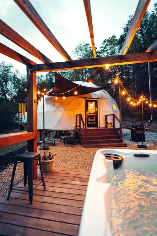 A cozy geodesic dome with string lights, a hot tub, and a wooden deck surrounded by trees at dusk.