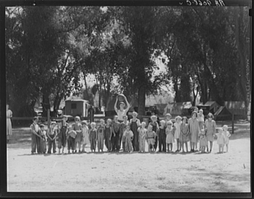 A large group of children stands outdoors, surrounded by trees and tents, smiling and posing for the camera.