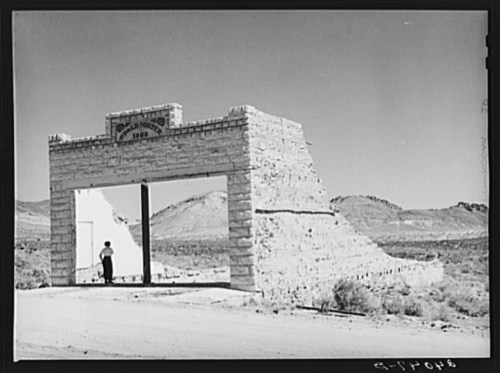 A stone archway with a sign reading "Goldfield, Nevada 1906" in a desert landscape, with a person standing nearby.