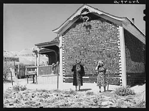 Two women stand in front of a house made of bottle bricks, with a desert landscape in the background.