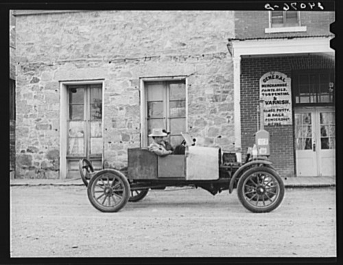 A vintage car parked in front of a stone building with a general store sign, featuring a man sitting inside the vehicle.
