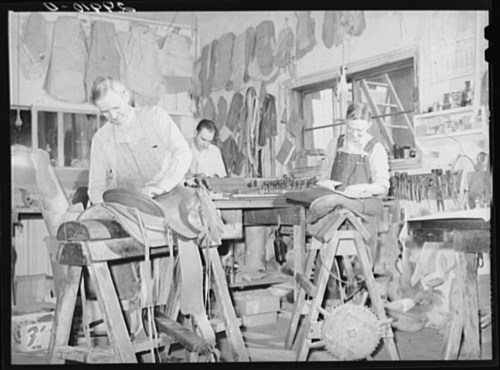 Three shoemakers work in a workshop, crafting shoes at wooden benches surrounded by tools and leather materials.