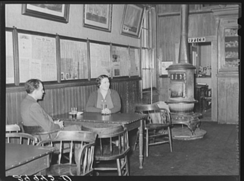 Two people sit at a table in a vintage bar, with wooden walls and a stove in the background.