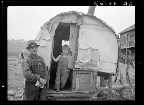 A man and a child stand by a weathered wagon in a rural setting, with mountains in the background.