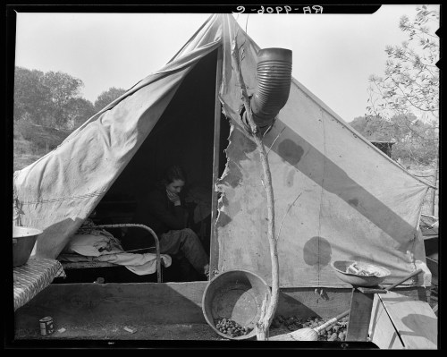 A person sits inside a tattered tent, surrounded by simple furnishings and outdoor scenery.