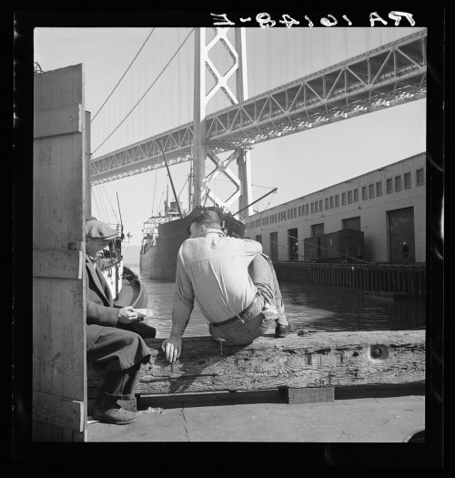 Two men sit on a dock near a ship, with the Bay Bridge in the background. The scene captures a moment of leisure by the water.