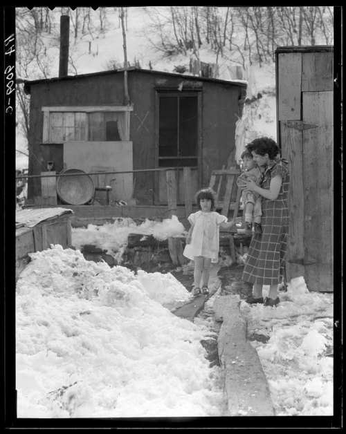 A woman holds a child while another child walks on a wooden path through snow, with a small cabin in the background.