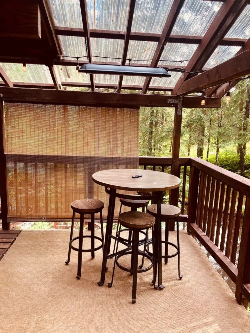 A wooden deck with a round table and four stools, surrounded by trees and covered by a transparent roof.