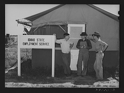 Three men stand outside a tent labeled "Idaho State Employment Service," with a rural landscape in the background.