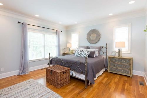 A cozy bedroom with a gray bedspread, wooden floors, and natural light from large windows. Decor includes a mirror and lamps.