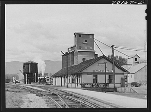 Historic railway station with grain silos and water tower, set against a cloudy sky and distant mountains.