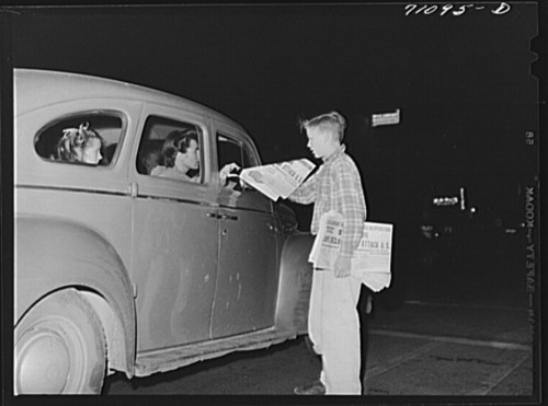 A boy sells newspapers to two girls in a car at night, illuminated by streetlights.