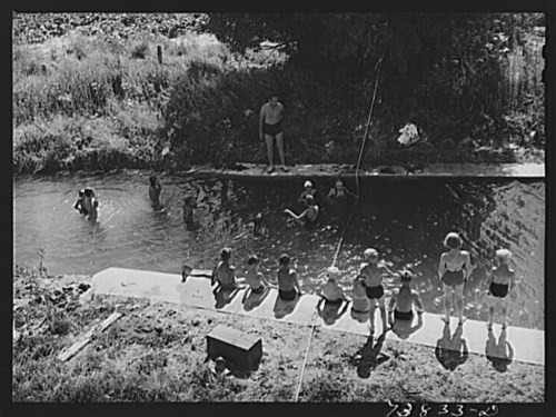 A group of children swimming and playing in a natural pool, with some sitting on the edge and others in the water.
