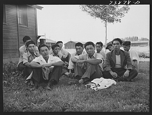A group of young men sitting on grass near a wooden building, with a few trees and structures in the background.