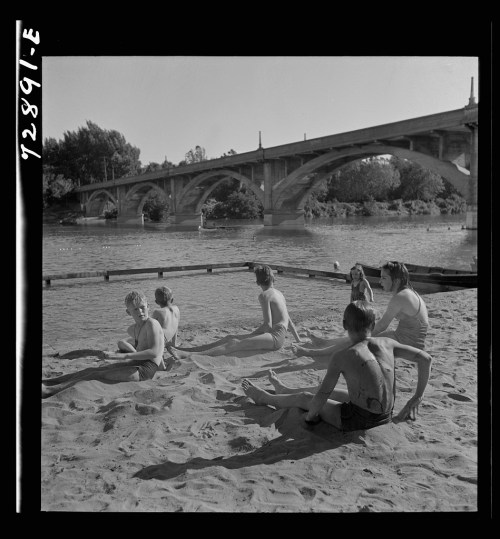 A group of children sitting on the sand by a river, with a bridge in the background and trees lining the shore.