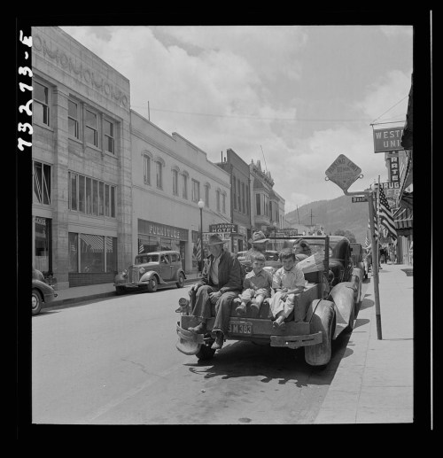 A vintage black-and-white photo of a family sitting on the back of a truck in a small town street.